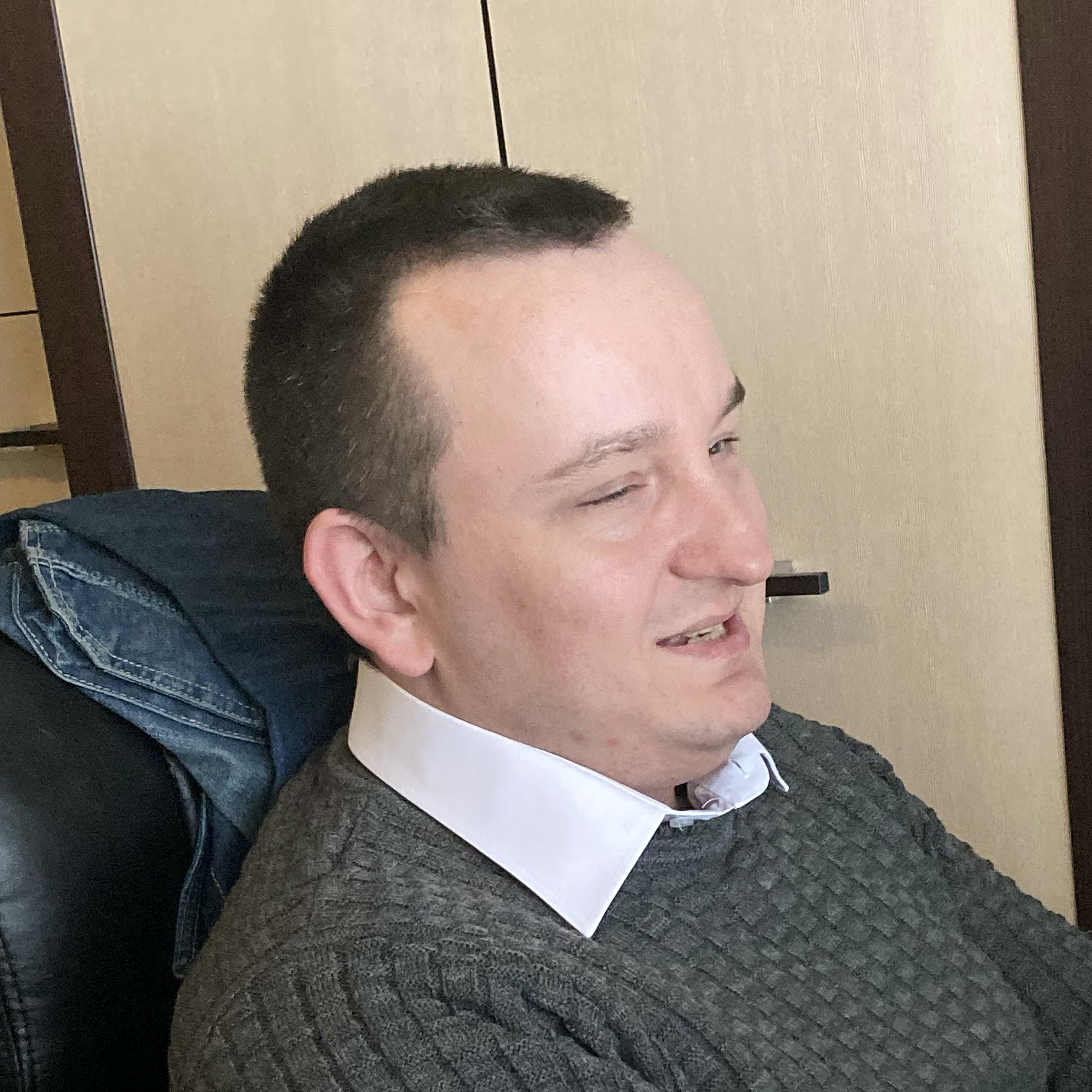 Maciej uśmiecha się ciepło, jakby rozmawiał z kimś obok. Ma ciemne, krótko ścięte włosy. Nosi szary sweter, spod którego widać kołnierzyk białej koszuli.