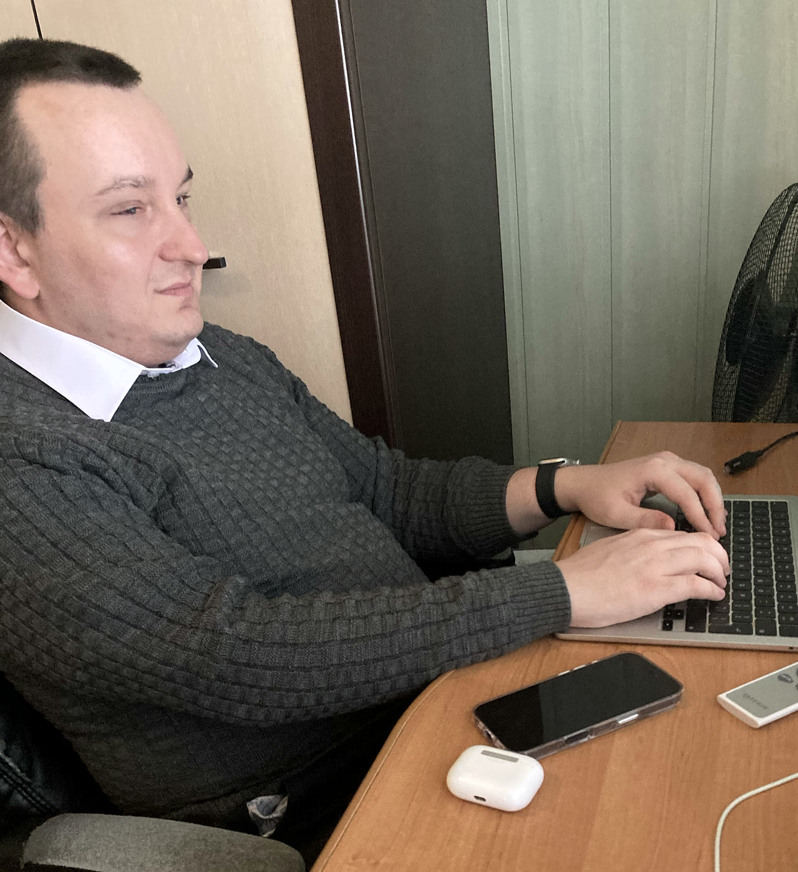 Maciej Walczak przy biurku pisze na srebrnym MacBooku. Przed laptopem leżą iPhone w przezroczystym etui i białe etui słuchawek AirPods. Na nadgarstku ma srebrny zegarek Apple Watch z czarną opaską Solo.
